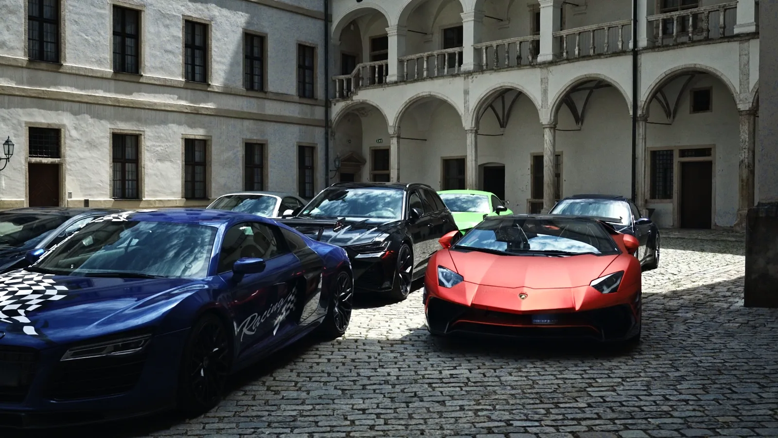 Mehrere Sportwagen in einem Arkadenhof auf Kopfsteinpflaster, vorn ein roter Lamborghini und ein blauer Audi R8