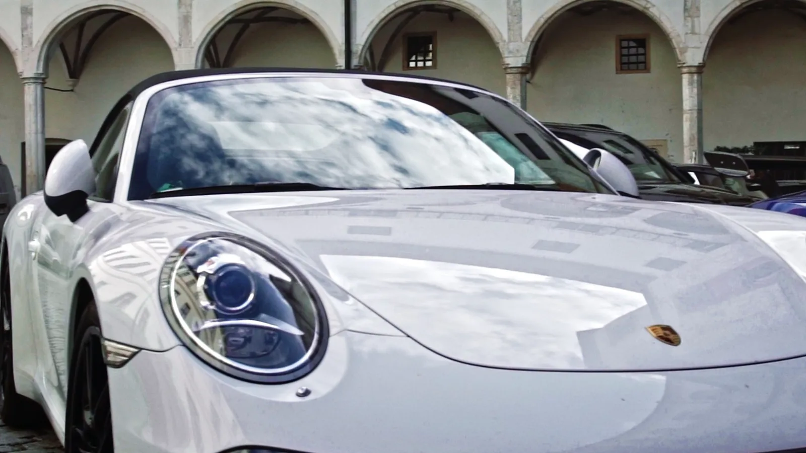 Front eines weißen Porsche 911, in Haube und Scheibe spiegeln sich Arkaden und Wolken