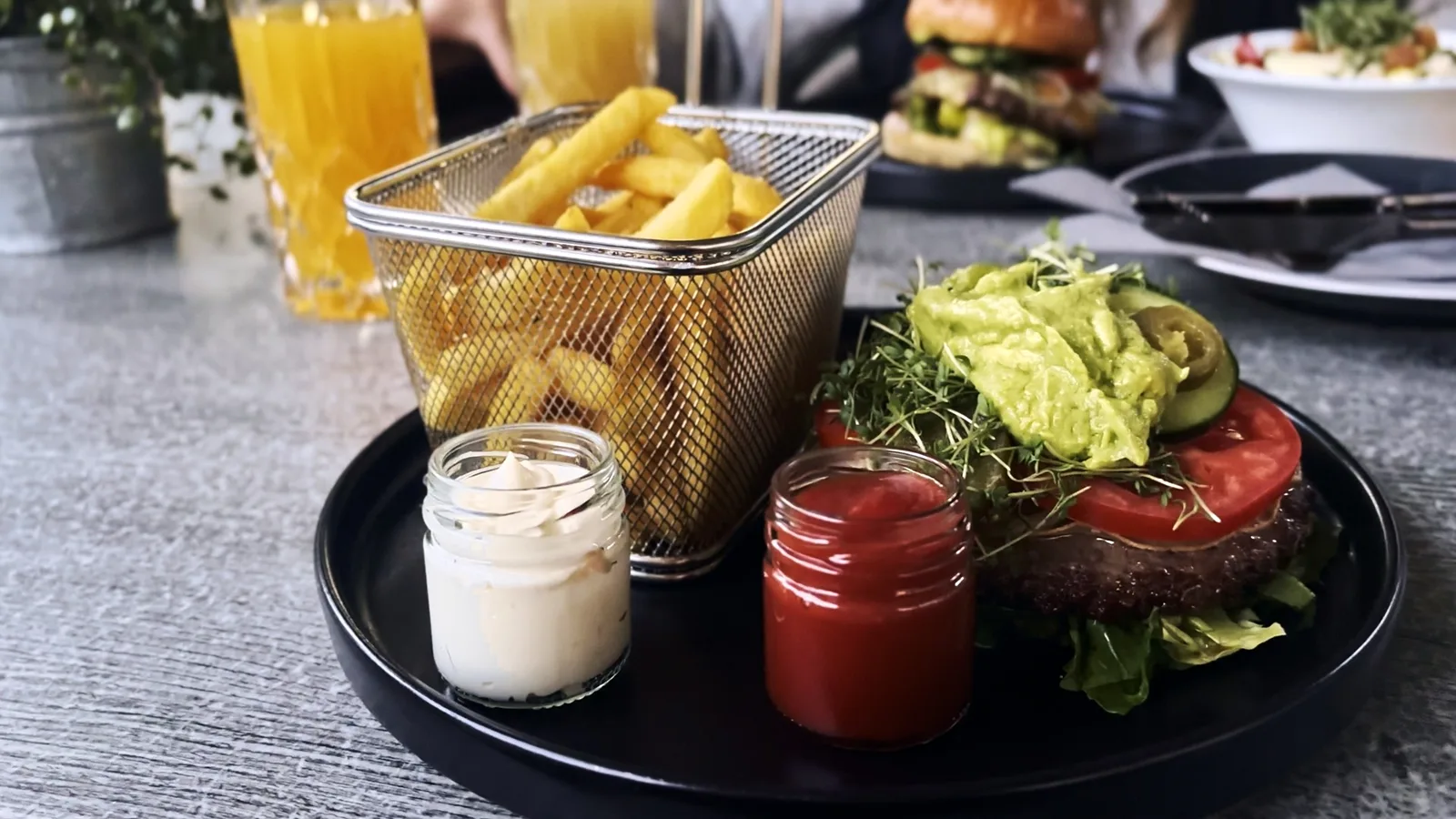 Burger mit Avocadocreme, Pommes im Drahtkorb und zwei Dips auf einem schwarzen Teller