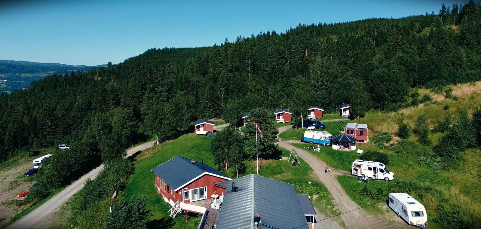 Campingplatz an einem Waldhang mit roten Hütten, Wohnmobilen und einem Wohnwagen, von oben gesehen