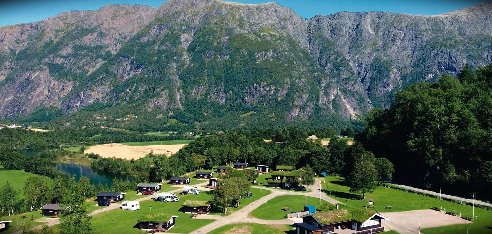 Campingplatz mit dunklen Holzhütten und Wohnmobilen auf grünen Wiesen, dahinter ein Fluss und eine hohe Felswand