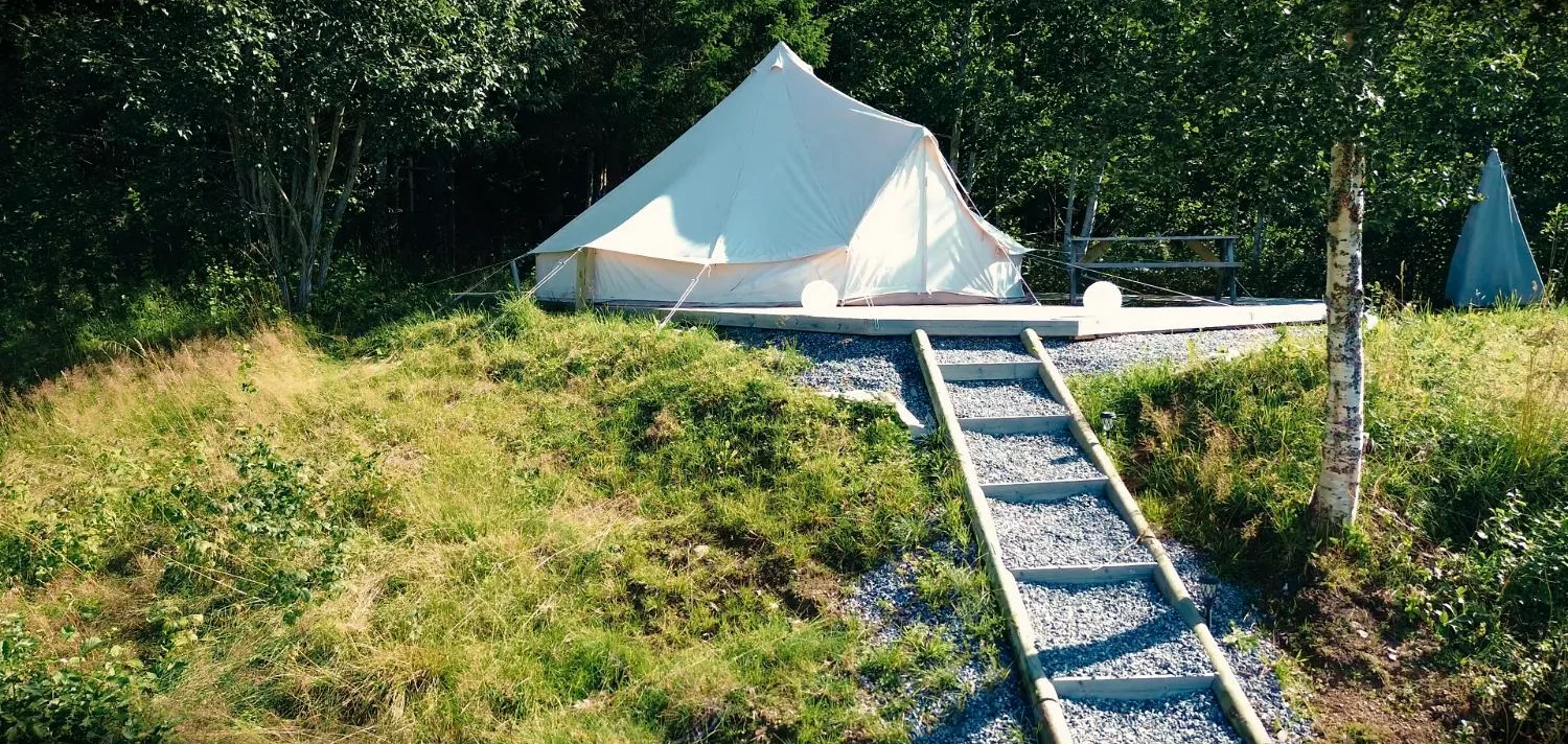 Großes helles Rundzelt auf einem Holzpodest am Waldrand, davor eine Treppe aus Kies und Holz