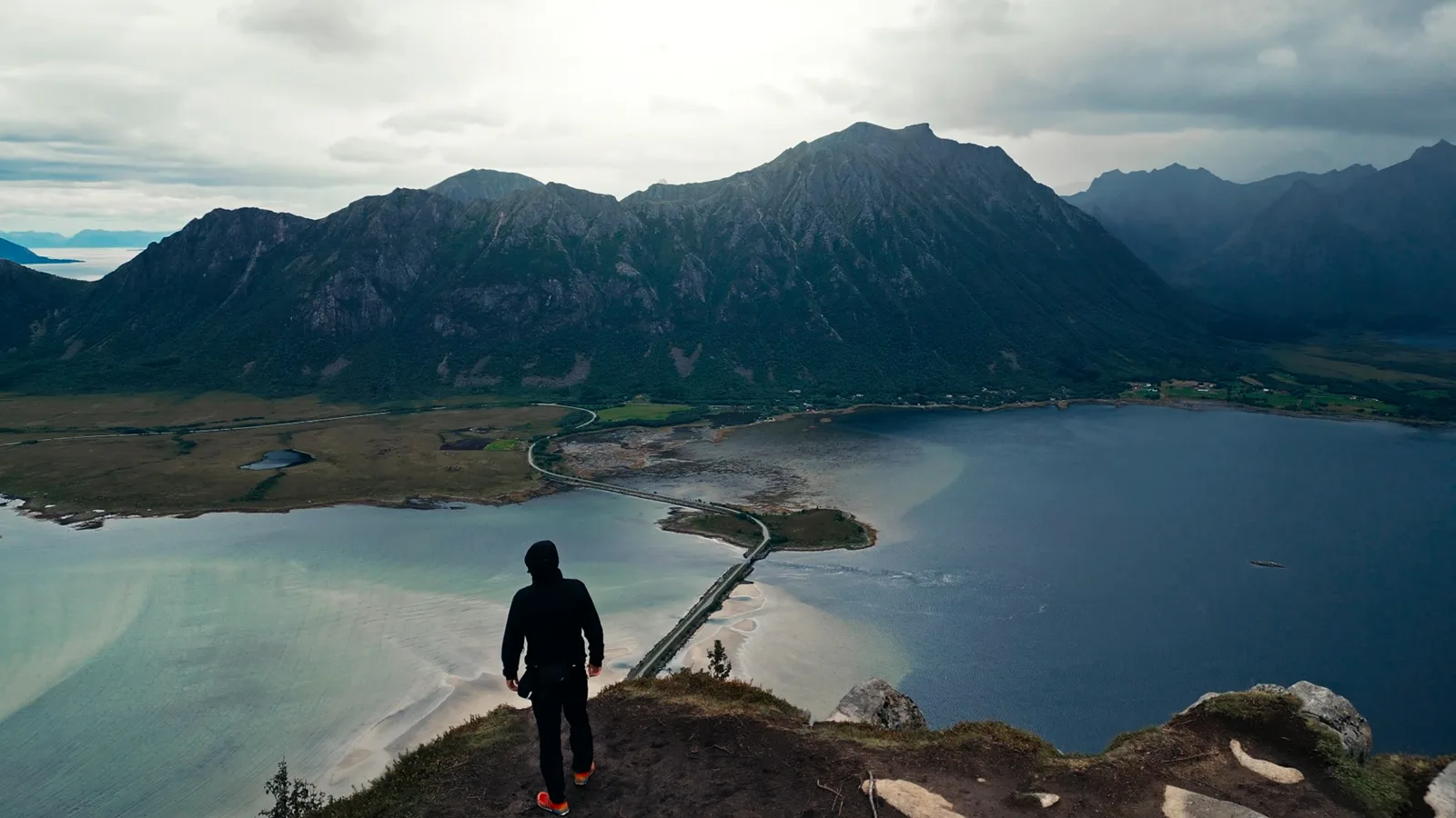 Person in dunkler Jacke steht mit dem Rücken zur Kamera auf einem Felsvorsprung über einer Fjordlandschaft, unten führt eine Straße über einen Damm