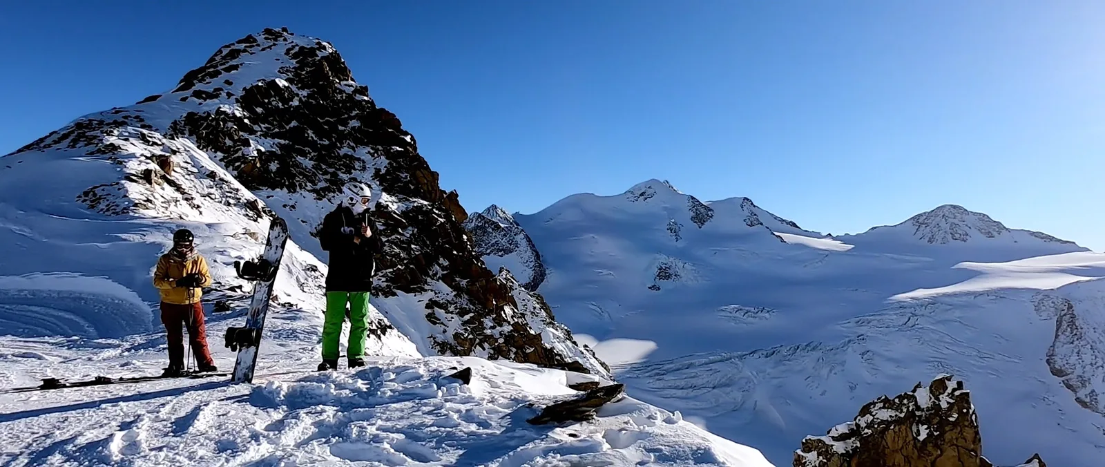 Zwei Wintersportler mit Snowboard auf einem Grat, dahinter ein weites Schneefeld