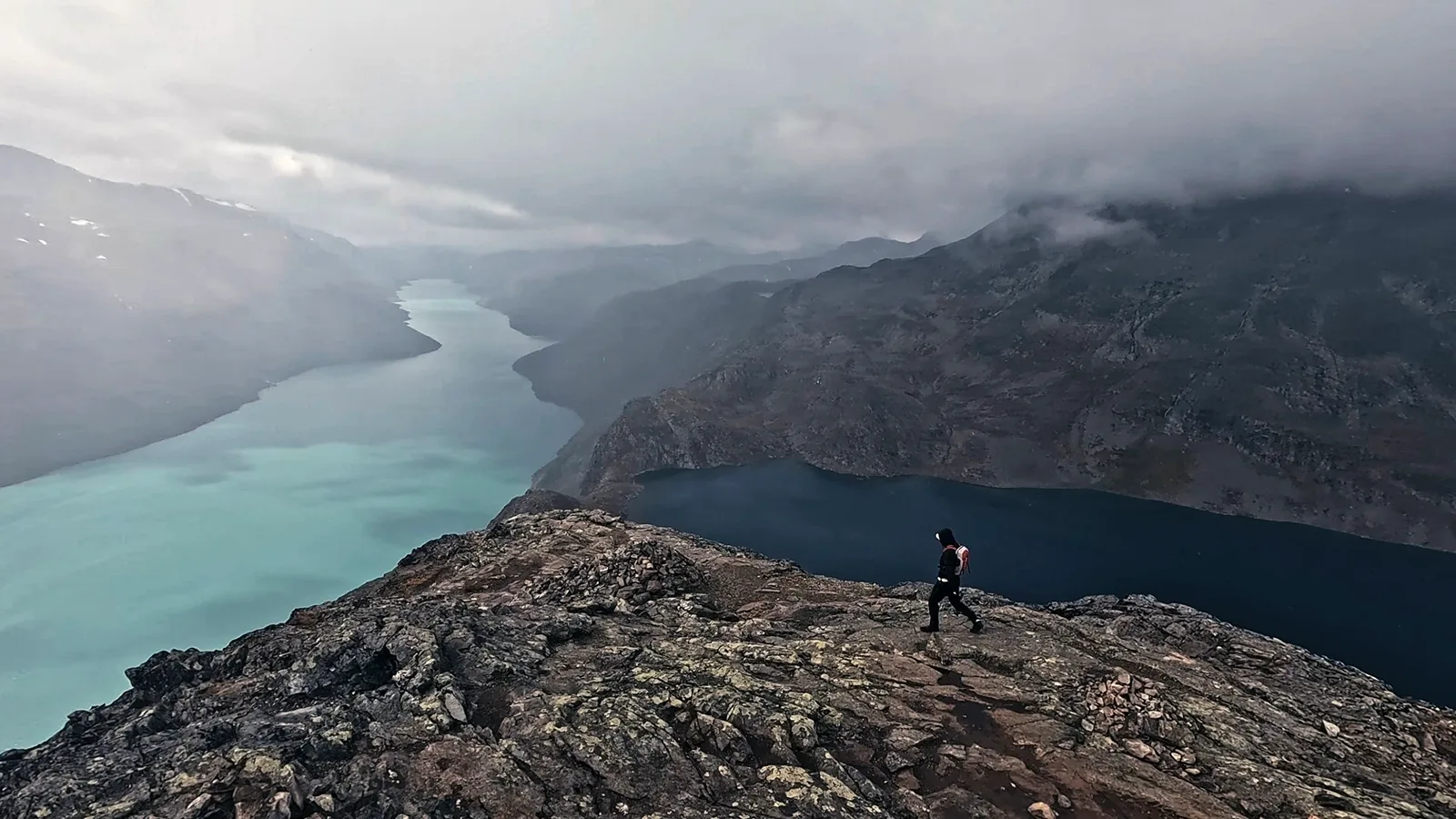 Wanderer mit Rucksack auf einem Felsgrat zwischen einem türkisfarbenen und einem dunkelblauen See, darüber tief hängende Wolken