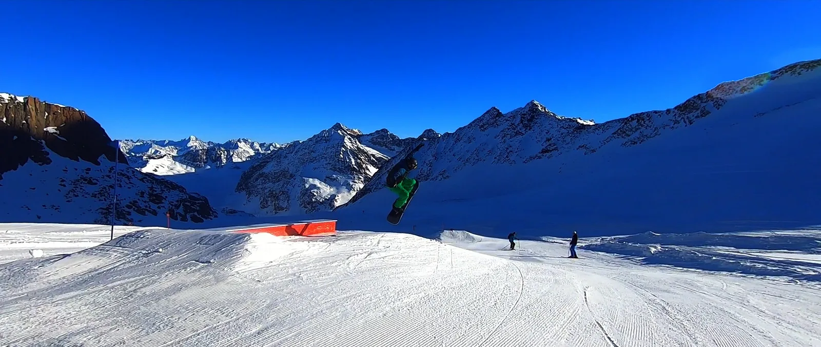 Snowboarder im Sprung über einem Funpark, im Hintergrund Berge