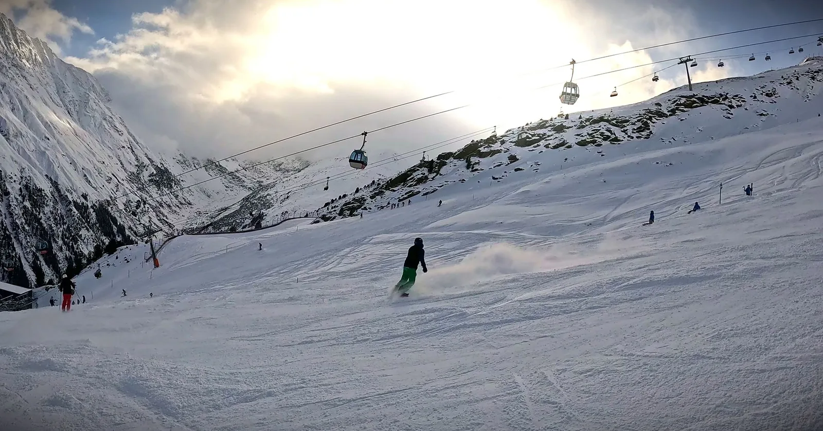 Snowboarder wirbelt Schnee auf, darüber Gondeln im Gegenlicht