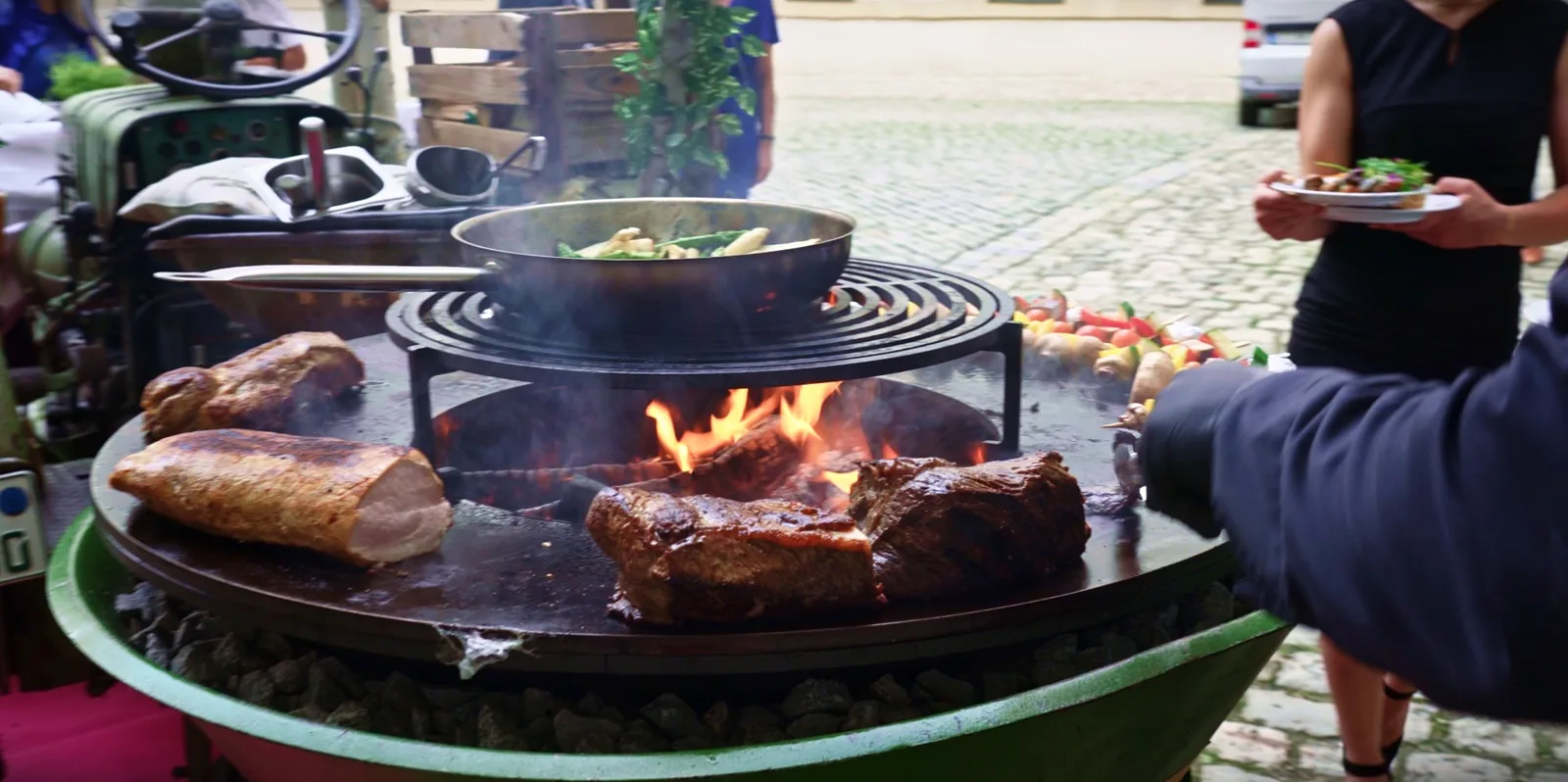 Fleisch und eine Gemüsepfanne auf einem Feuerring, im Hintergrund Gäste mit Tellern