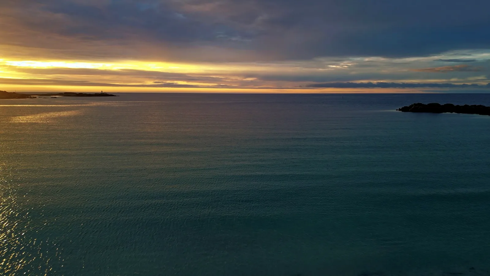 Ruhiges Meer am Abend, über dem Horizont ein gelber Lichtstreifen unter einer Wolkendecke, rechts eine Felsinsel
