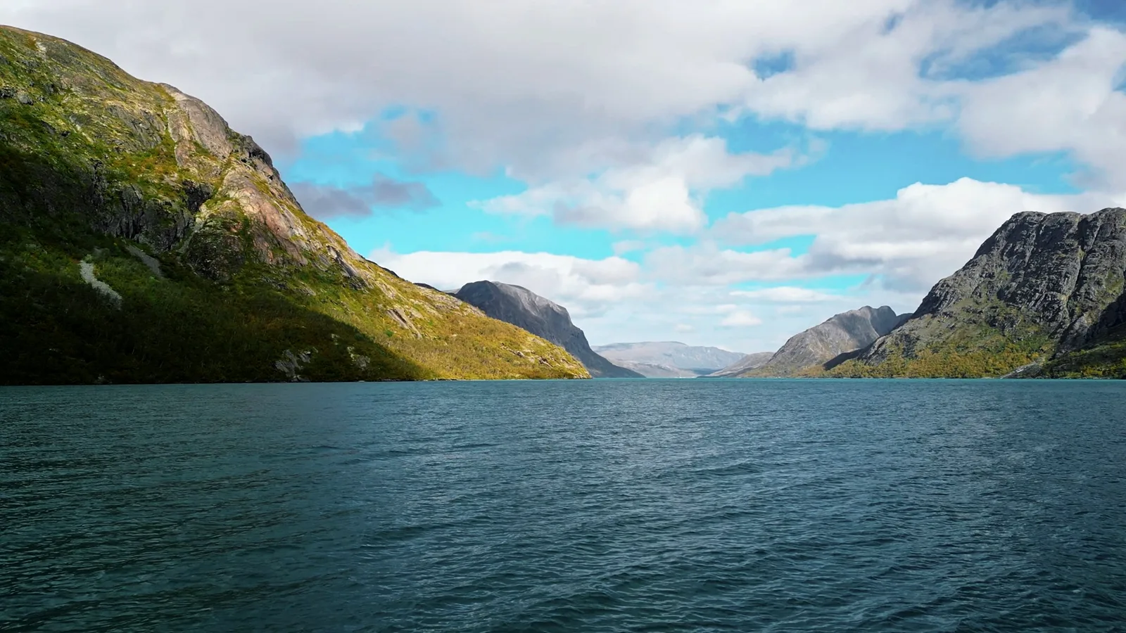 Weiter Bergsee zwischen steilen Hängen, darüber aufgelockerte Wolken