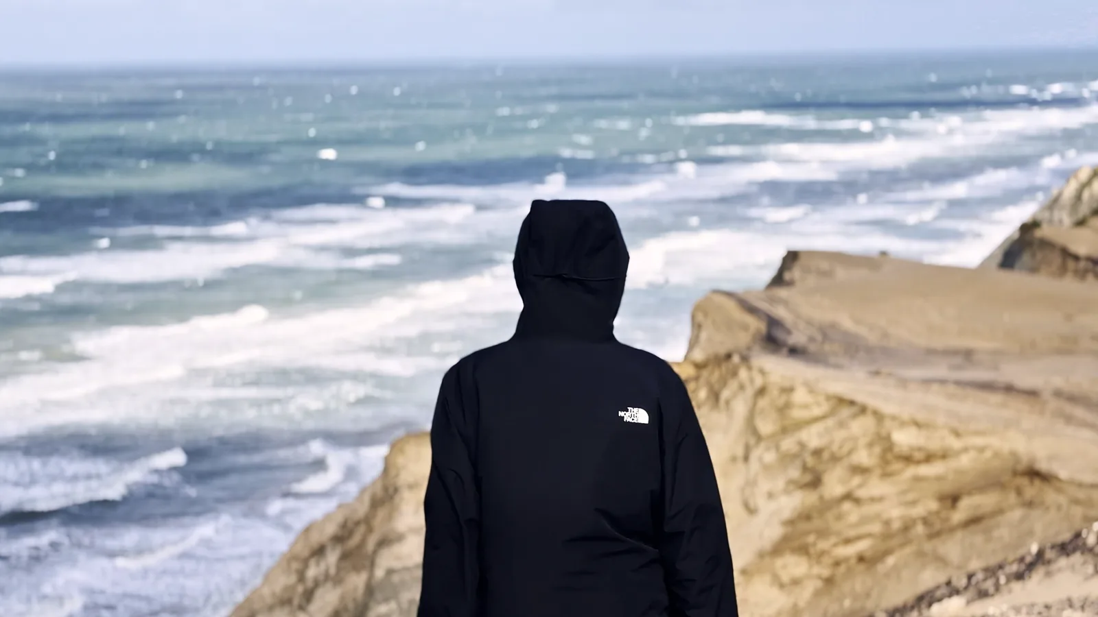 Person mit Kapuze blickt von einer Klippe auf die Brandung