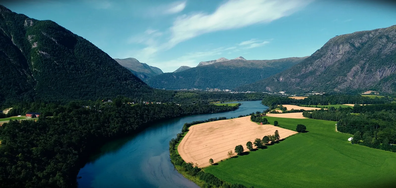 Fluss windet sich durch ein Tal mit Kornfeldern und Wiesen, links und rechts bewaldete Berge