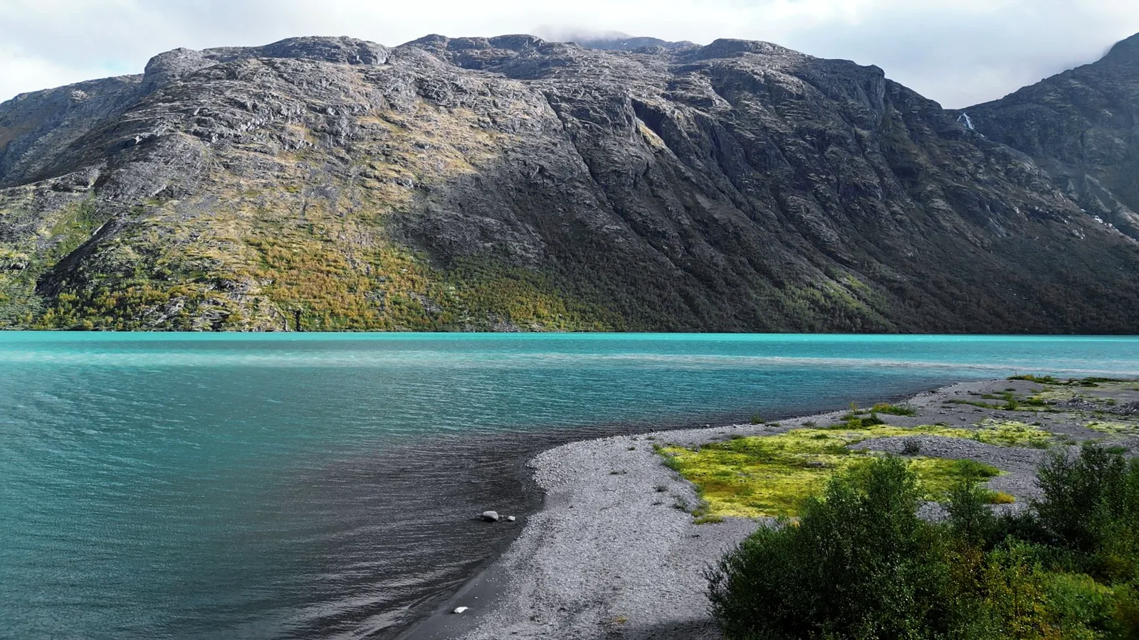 Türkisfarbener Bergsee mit Kiesufer und Moospolstern, dahinter eine graue Felswand mit herbstlich gefärbten Birken