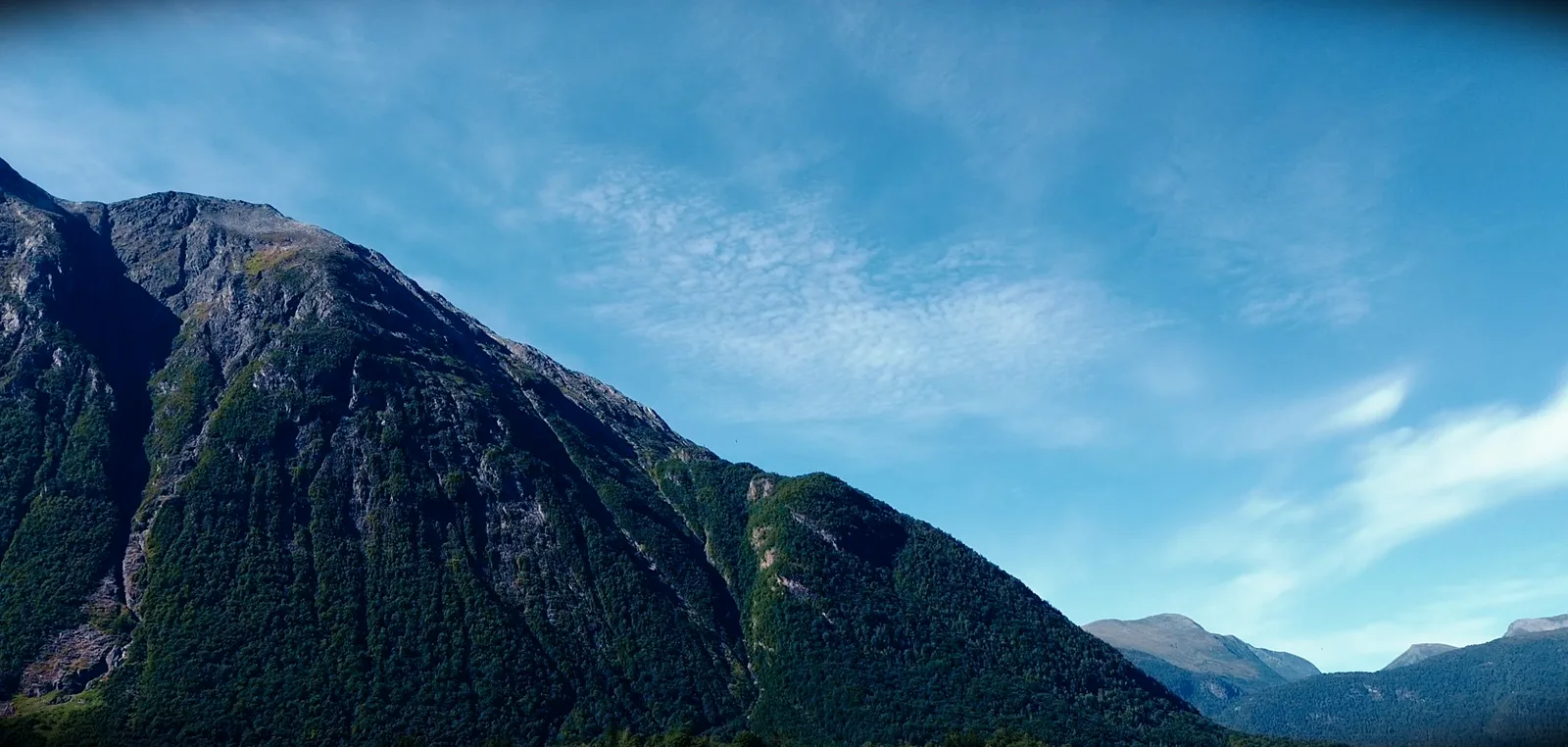 Steiler bewaldeter Berghang vor blauem Himmel mit feinen Schäfchenwolken