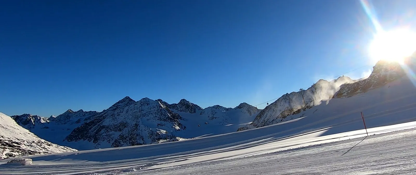 Verschneite Gipfel im Schatten, am Bildrand die tief stehende Sonne über einer Piste
