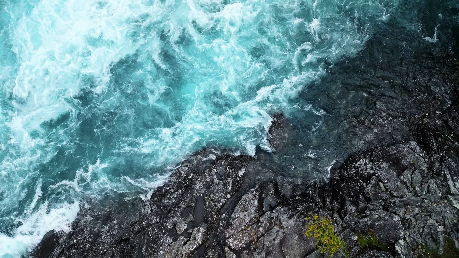 Schäumendes türkisfarbenes Wasser trifft auf dunklen Fels, senkrecht von oben gesehen